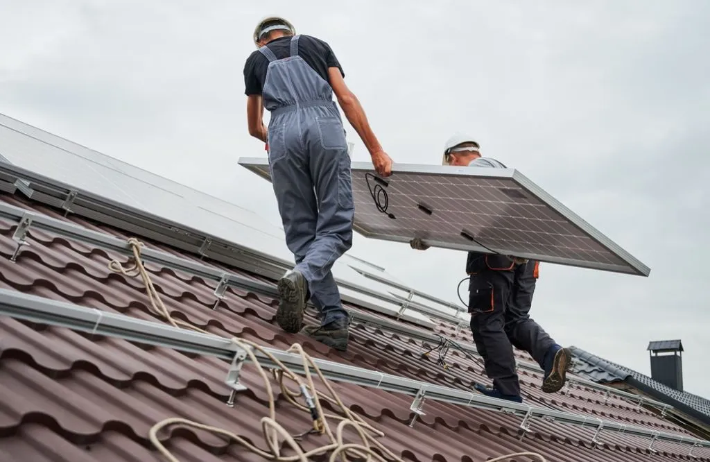 The Most Detailed Review of Tesla Powerwall: Effectivity, Pros and Cons 4 Men technicians carrying photovoltaic solar panels on the roof of the house.