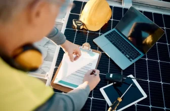 Solar engineer writing notes near solar panels, laptop, and his helmet.